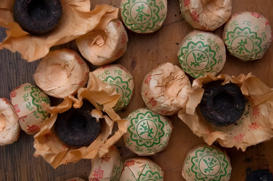 Several round tea cakes wrapped in decorative paper with green and red designs, partially unwrapped to reveal pu-erh tea inside, placed on a wooden surface.