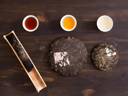 a dark wooden surface with two pu-erh tea cakes, one dark and one lighter, and a bamboo container with pu-erh tea.